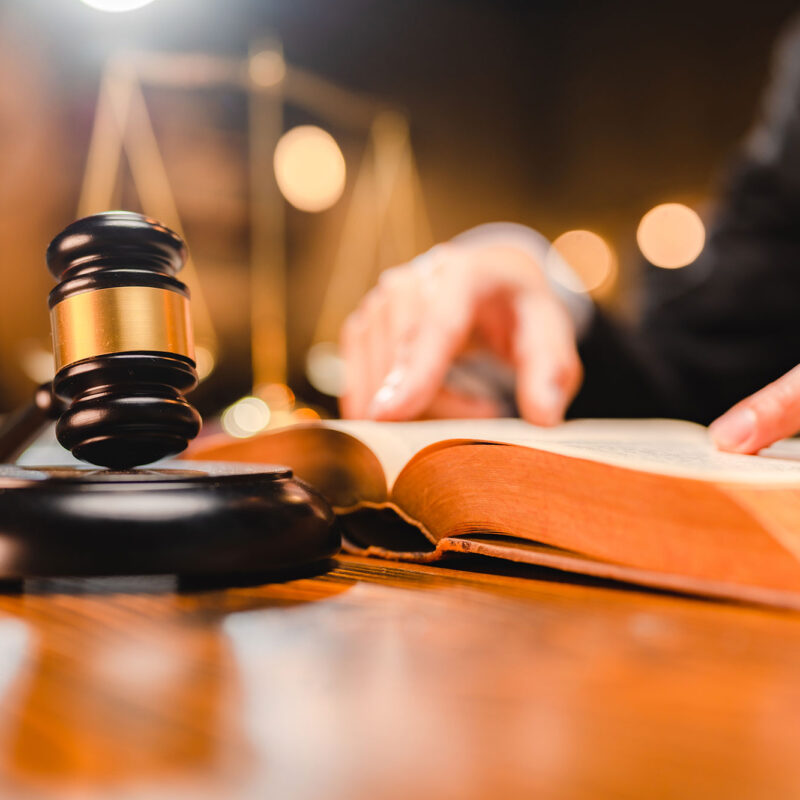 judges gavel on desk with man reading law book in background